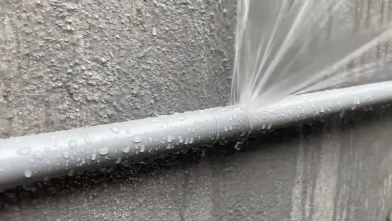 Water spraying forcefully from a burst plastic pipe mounted on a wall, showing a severe plumbing emergency